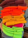 A person in a store holds a stack of colorful knit beanie hats in shades of burgundy, orange, yellow, pink, and neon green.