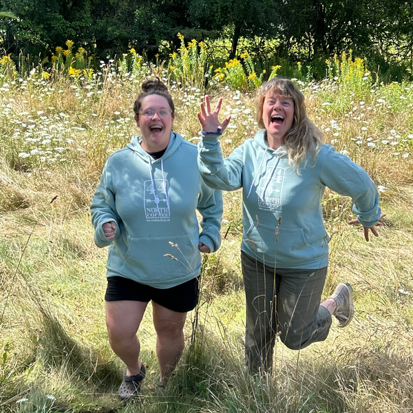 Two people in matching DUSTY blue hoodies running through a field with wildflowers and trees.