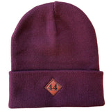 A dark plum purple knit beanie hat with a brown leather diamond 44 North Coffee patch on the cuff laid flat on a white background