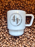 A mist blue milk glass coffee mug with a black circular logo reading '44 NORTH COFFEE', placed on a bed of roasted coffee beans. The mug features a stacked design with a wide handle and a thick base.
