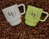Two milk glass coffee mugs with a stacked design, one mist blue and one jade green, both printed with a black circular logo reading '44 NORTH COFFEE'. They are arranged on a bed of roasted coffee beans, with the mist blue mug on the left and the jade green mug on the right.