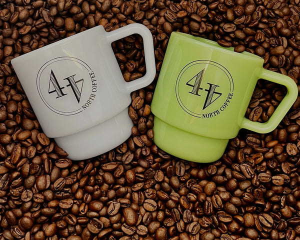 Two milk glass coffee mugs with a stacked design, one mist blue and one jade green, both printed with a black circular logo reading '44 NORTH COFFEE'. They are arranged on a bed of roasted coffee beans, with the mist blue mug on the left and the jade green mug on the right.